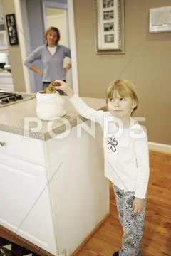 A mother watching her daughter take a cookie from a jar on the kitchen counter Stock Photos