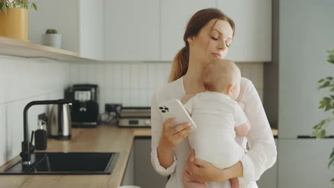 Mother is working on the phone looking back at the baby she is holding in her Stock Footage 208908279