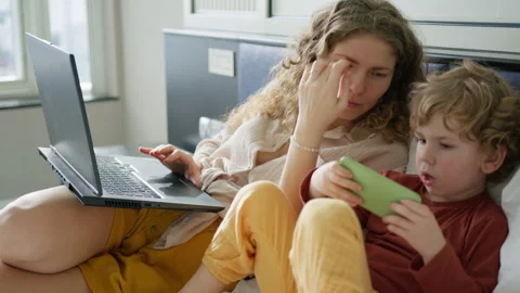 Mother works on her laptop while her child plays on a smartphone, showcasing Stock Footage 296513484
