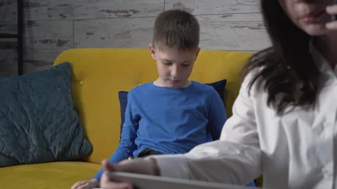 Mother works while sitting on the couch with her son, who is looking at a tablet 動画素材 259309293