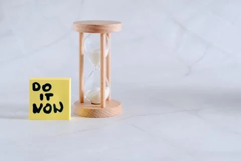 A motivational yellow sticky note DO IT NOW next to an hourglass on a desk 写真素材