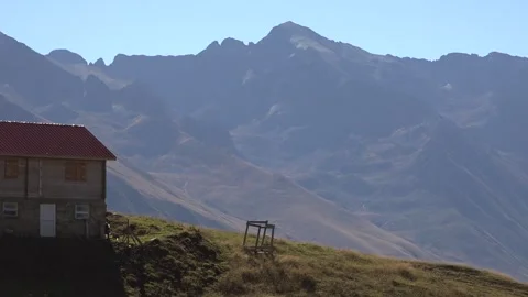 Mountain cabin stands on grassy meadow ridge below snowy peaks and clear sky Stock Footage 339477103