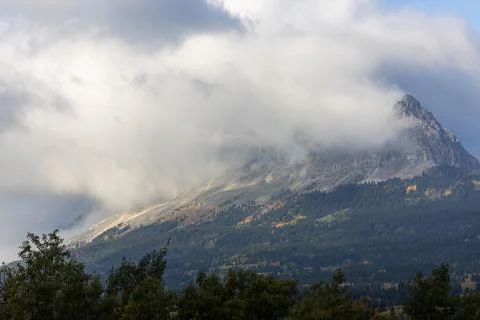 A mountain covered in clouds with a few trees in the foreground Stock Photos