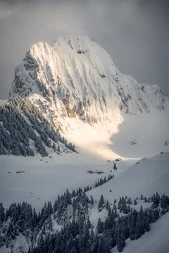 A mountain covered with snow on which the sunlight falls next to a house stan Stock Photos
