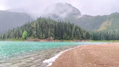 Mountain lake surrounded by pine forest and misty peaks under cloudy sky Stock Footage 340533337