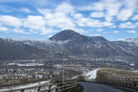 Mountain landscape under cloudy sky with winding road and snow-covered city Stock Photos