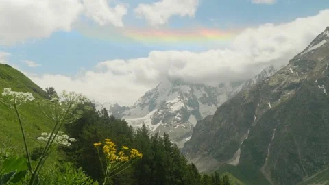 Mountain peaks covered with snow woth thick fluffy clouds and a rainbow above Stock Footage 138743376