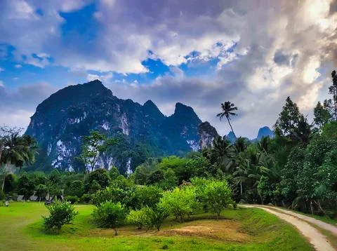 Mountains and trees line a path in a rural area with clouds in the sky duri.. Stock Photos
