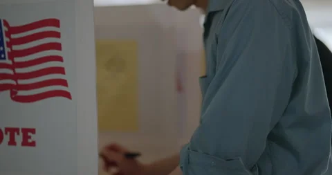 Move from backs to profile of voters at election booths, US flag behind Video stock 275539964