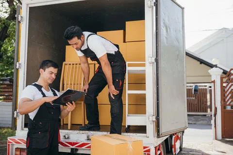 Mover workers unload boxes inspecting shipment with a clipboard near the truck Stock Photos