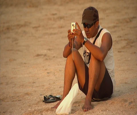 MS man sitting on beach sand, looking through camera Stock Footage 86747463
