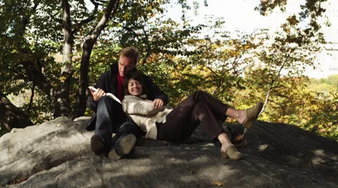 MS PAN Man reading book to woman resting on his lap in Central Park, New York Vídeos de archivo 38803529