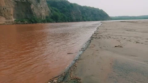 Muddy River Flowing Beside Sandy Bank And Steep Rocky Cliffs Stock Footage 345943809