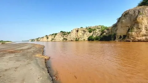 Muddy River Flowing Past Massive Rocky Cliffs Under Blue Sky 스톡 사진