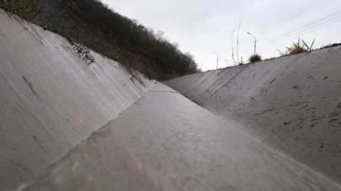 Muddy water flows through a concrete drainage ditch next to a road Видео 306974978