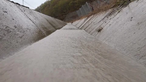 Muddy water flows through a concrete drainage ditch next to a metal fence Vidéo 306974987