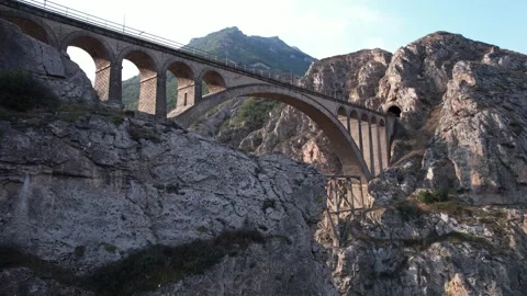Multi arch concrete bridge spans deep valley with mountain backdrop under Stock Footage 342704100