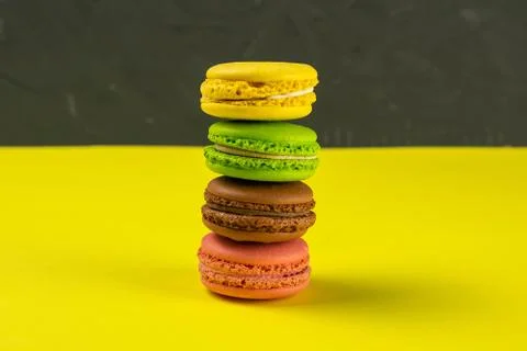 Multi-colored macaroons stand in a pile on a yellow table near a black backgr Stock Photos