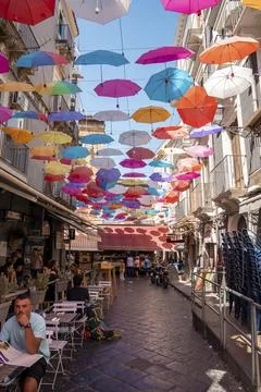 Multi colored umbrellas hanging over tourists relaxing at cafe amidst buildings Stock Photos