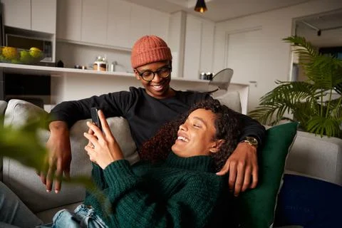 Multi-ethnic couple looking at each other, laughing about text on phone. On sofa Stock Photos