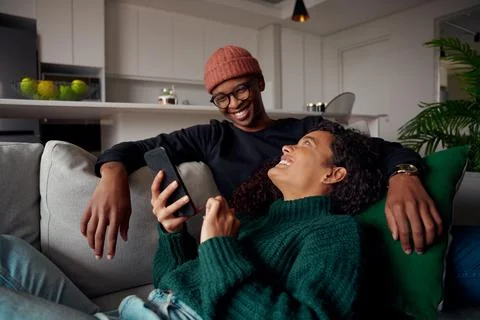 Multi-ethnic couple looking at each other, laughing about text on phone. On sofa Stock Photos
