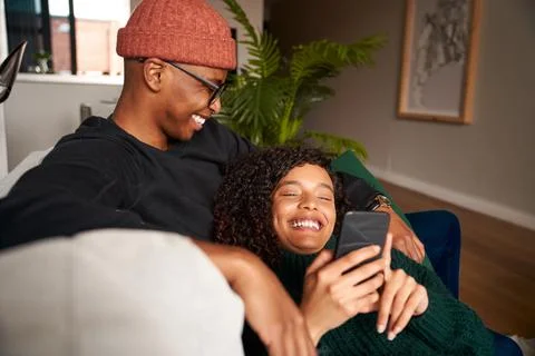 Multi-ethnic couple smiling while looking at cellphone. Modern apartment on the Stock Photos