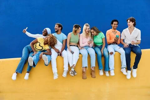 Multi-ethnic young people using phone sitting on a colorful wall Stock Photos