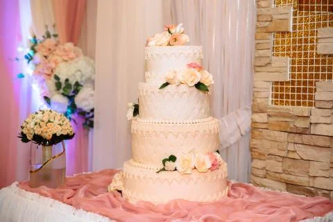 A multi-level white wedding cake decorated with flowers stands on a table next Stock Photos