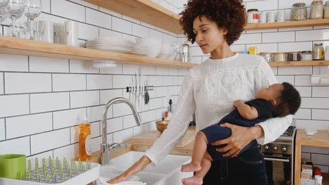 Multi-Tasking Mother Holds Sleeping Baby Son Whilst Cleaning And Working On Stock Footage