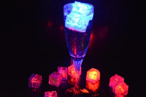 Multicolored glowing ice cubes float in champagne glass that illuminates drink. Stock Photos