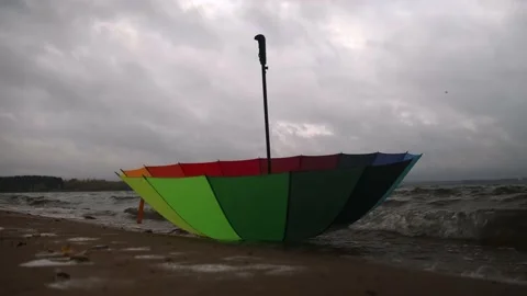 A multicolored umbrella washed ashore by waves, cloudy windy weather Stock Footage 219961863