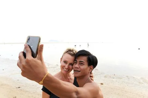 Multicultural couple embracing while taking a selfie on a sandy beach Stock Photos
