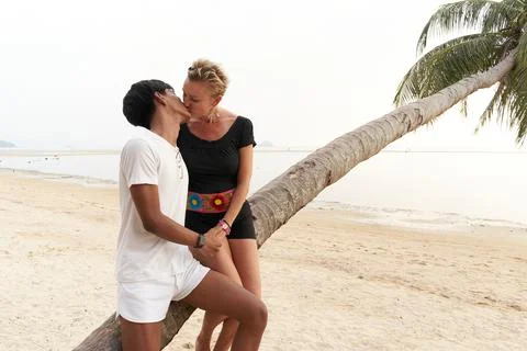 Multicultural lovers kissing while sitting on a tropical beach Stock Photos