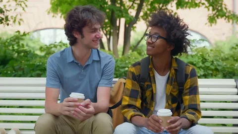 Multiethnic college students sitting on bench relaxing after classes drinking Stock Footage 235450823
