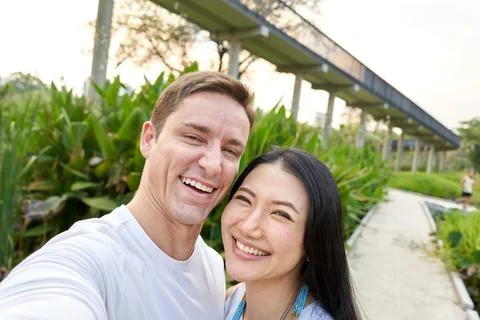 Multiethnic couple laughing while taking a selfie in an urban park during sunset Stock Photos