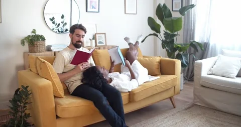 Multiethnic couple reading a book lying on the sofa at home in an apartment Stock Footage 172525576
