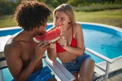 Multiethnic couple sitting by the pool eating together one slice of watermelo Stock Photos