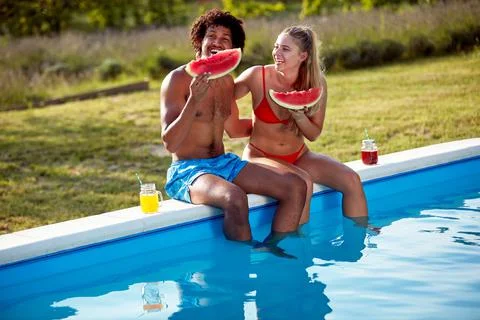 Multiethnic couple sitting on the side of the pool with legs in water, eating Stock Photos