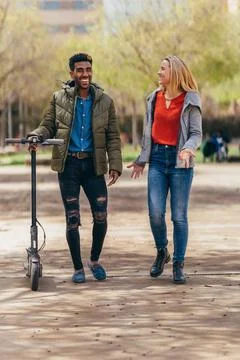 Multiethnic couple talking while walking, he is carrying an electric scooter Stock Photos