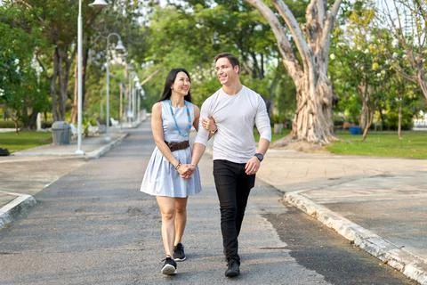Multiethnic couple walking together on a path in a city park during sunset Stock Photos