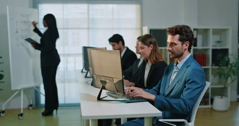 Multiethnic employees type at desktop computers as presenter writes on board 库存影片 348067751