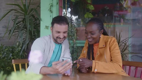 Multiethnic friends looking at mobile phone sitting outdoors at cafe Stock Footage 244546014