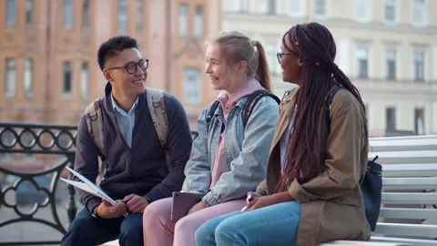Multiethnic friends talk sitting on bench in city. Realtime Stock Footage 233830702