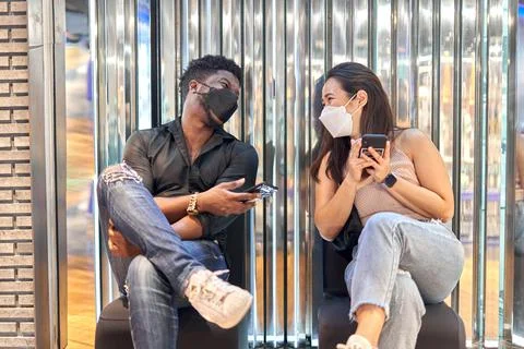 Multiethnic friends wearing mask using the mobile and sitting on a shopping mall Stock Photos