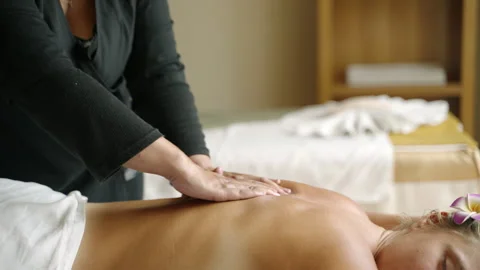 Multiethnic masseuse gently massages back of a young blonde causasian woman for 스톡 동영상 251113281