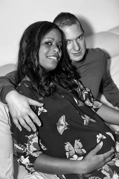 Multinational couple sitting on the couch with her pregnant Stock Photos