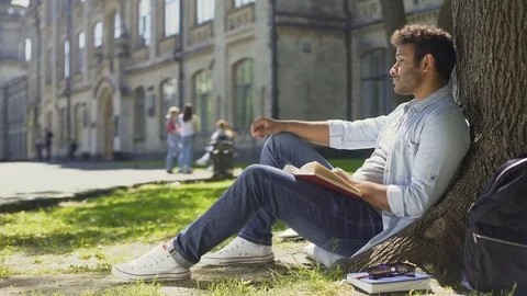 Multinational guy sitting under tree, reading book looking around, leisure time Stock Footage 77195605