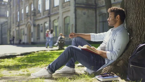 Multinational guy sitting under tree, reading book looking around, leisure time Stock Footage 87274734