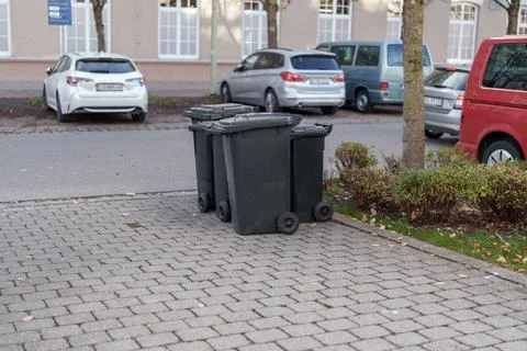 Multiple black trash bins lined up on a sidewalk near parked cars in a quie.. 스톡 사진