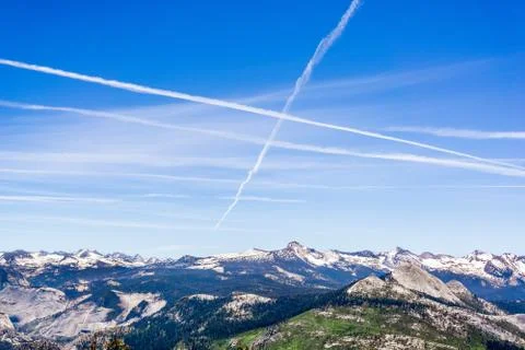 Multiple contrails cross paths through the blue sky above snow capped mountai Stock Photos